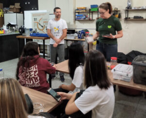 fotografia de uma sala exibindo 4 estudantes sentadas de costas e dois professores em pé, de frente, explicando os processos